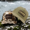 Olive Expedition Hat and Sportsman's Hat on a rock in front of a river.